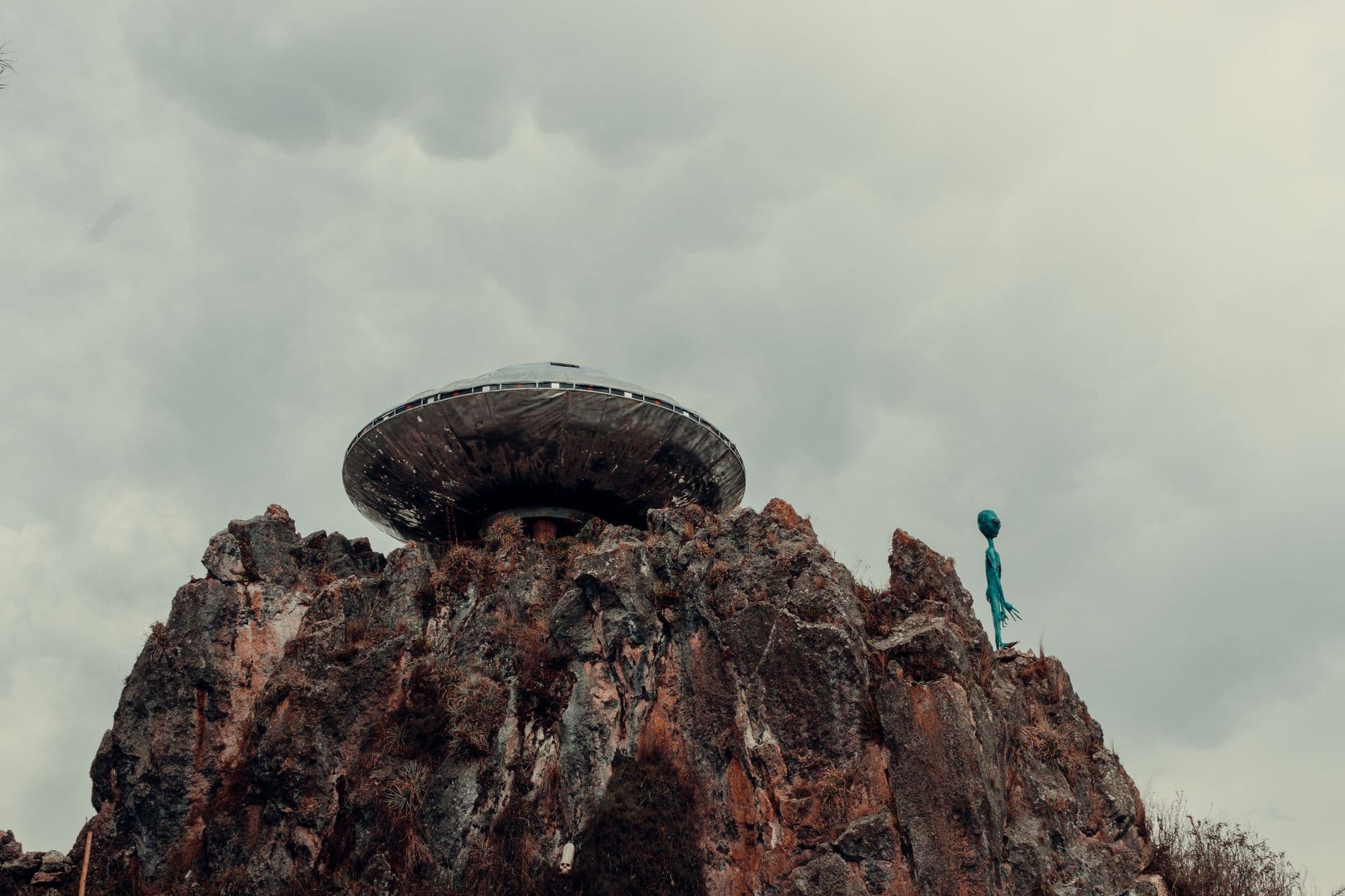 A silver disc-shaped craft on high ground under an overcast sky, with a thin dark figure standing to one side